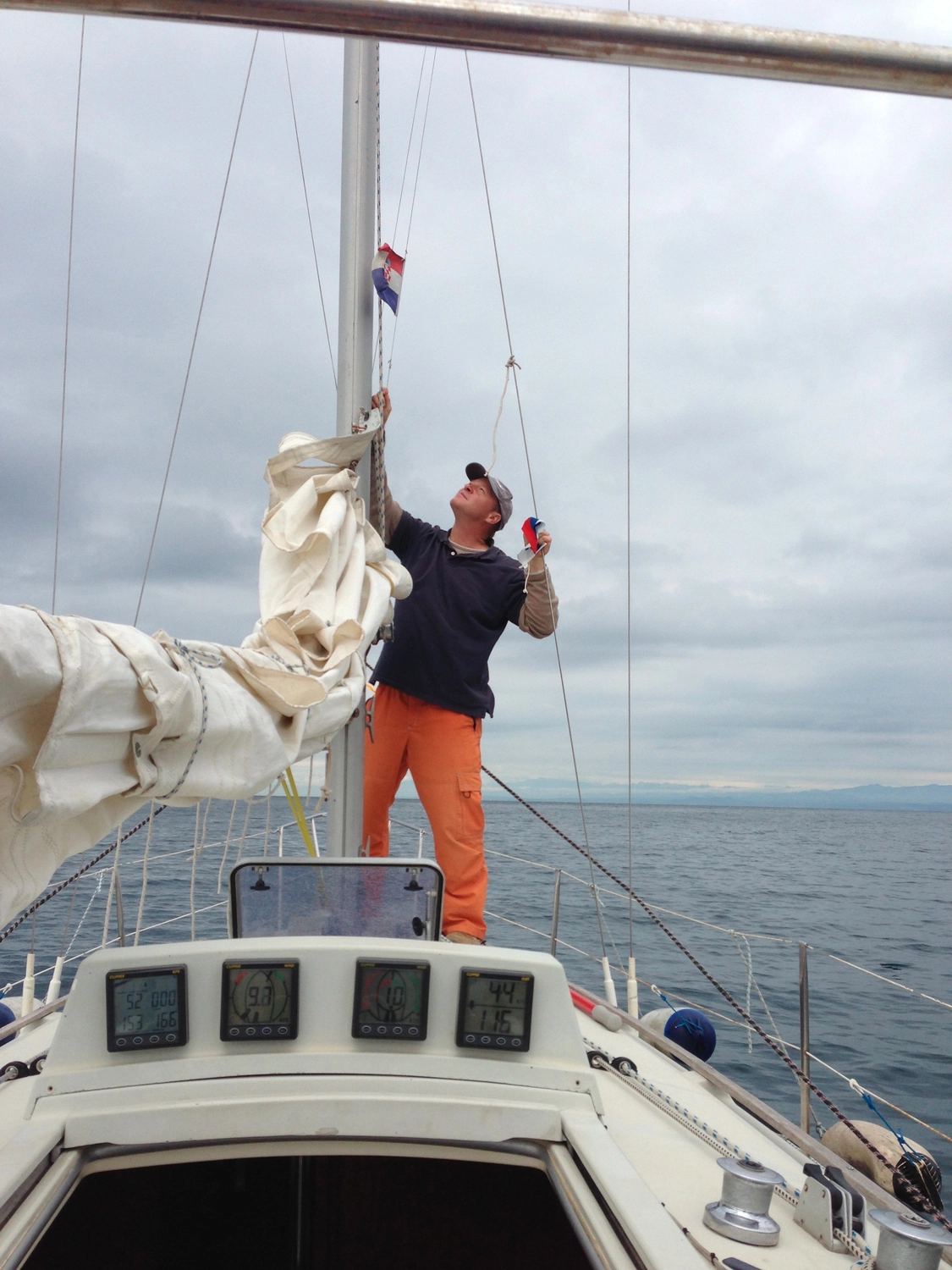 Motorboot-u. Segelschule Ravensburg - Wassersportausbildung und lizenzierung - Ein Mann in einem dunklen Hemd und einer orangefarbenen Hose steht an Deck eines Segelboots und justiert die weißen Segel vor einem bewölkten Himmel.