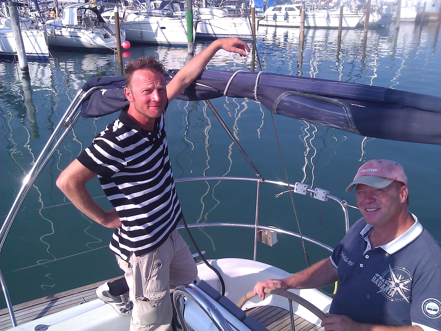 Motorboot-u. Segelschule Ravensburg - Wassersportausbildung und lizenzierung - Ein Mann in einem gestreiften Hemd steht auf einem Segelboot in einem Jachthafen, während ein anderer Mann am Steuer sitzt.