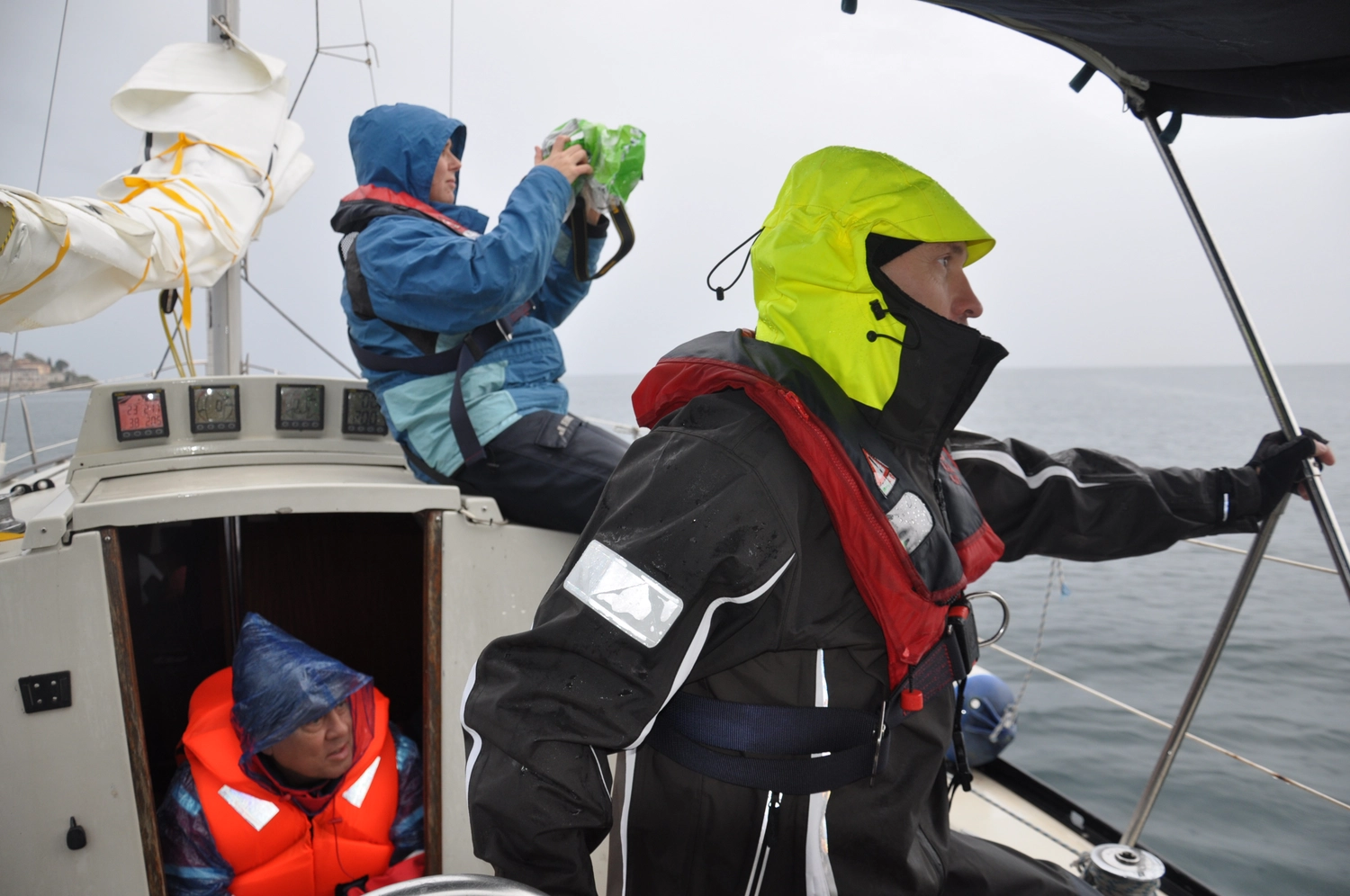 Motorboot-u. Segelschule Ravensburg - Wassersportausbildung und lizenzierung - Zwei Personen in voller Segelausrüstung auf einem Boot; eine steht im Vordergrund und trägt eine gelbe Kapuze und eine schwarze Jacke, während die andere im Hintergrund sitzt und eine blaue Jacke trägt.