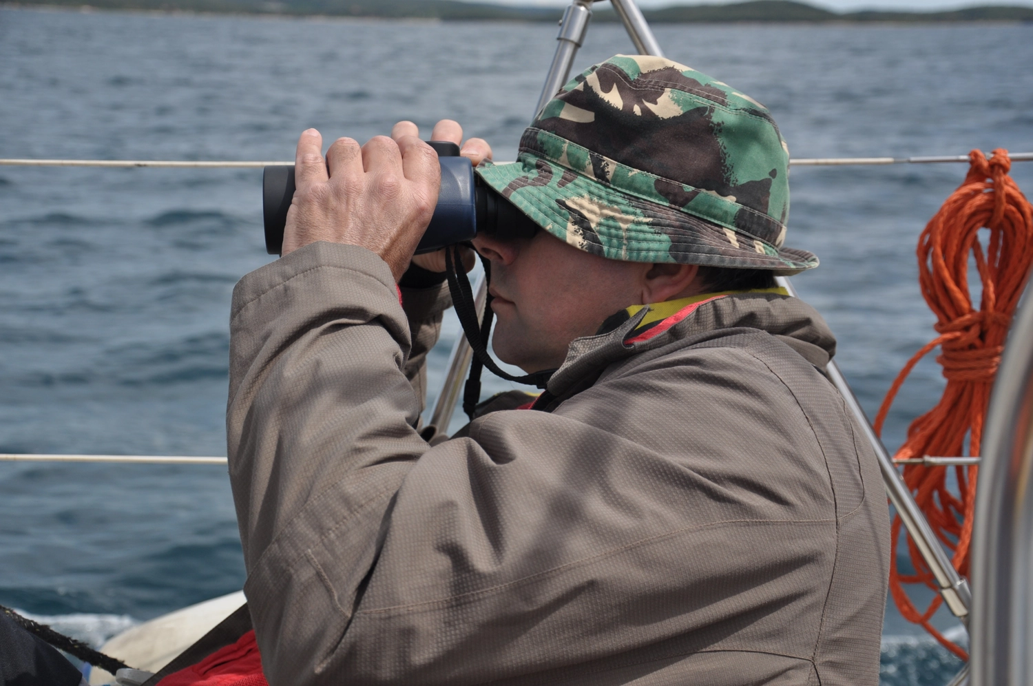 Motorboot-u. Segelschule Ravensburg - Wassersportausbildung und lizenzierung - Eine Person in Tarnmütze und -jacke schaut an Bord eines Schiffes durch ein Fernglas.