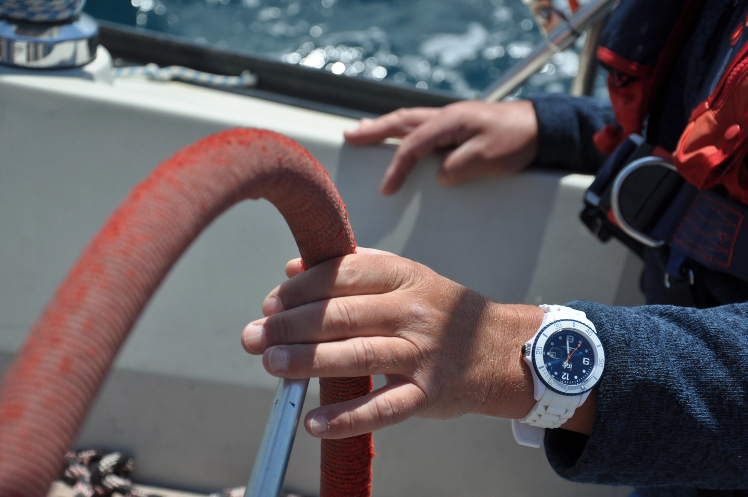 Motorboot-u. Segelschule Ravensburg - Wassersportausbildung und lizenzierung - Nahaufnahme der Hände einer Person an einem roten Steuerrad eines Bootes, an deren Handgelenk eine weiß-blaue Uhr zu sehen ist.