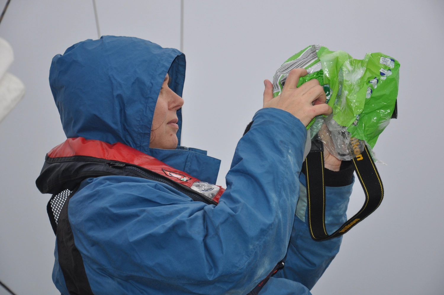 Motorboot-u. Segelschule Ravensburg - Wassersportausbildung und lizenzierung - Eine Person in einer blauen Kapuzenjacke und Schwimmweste hält eine Kamera, die von einer leuchtend grünen wasserdichten Hülle geschützt wird.