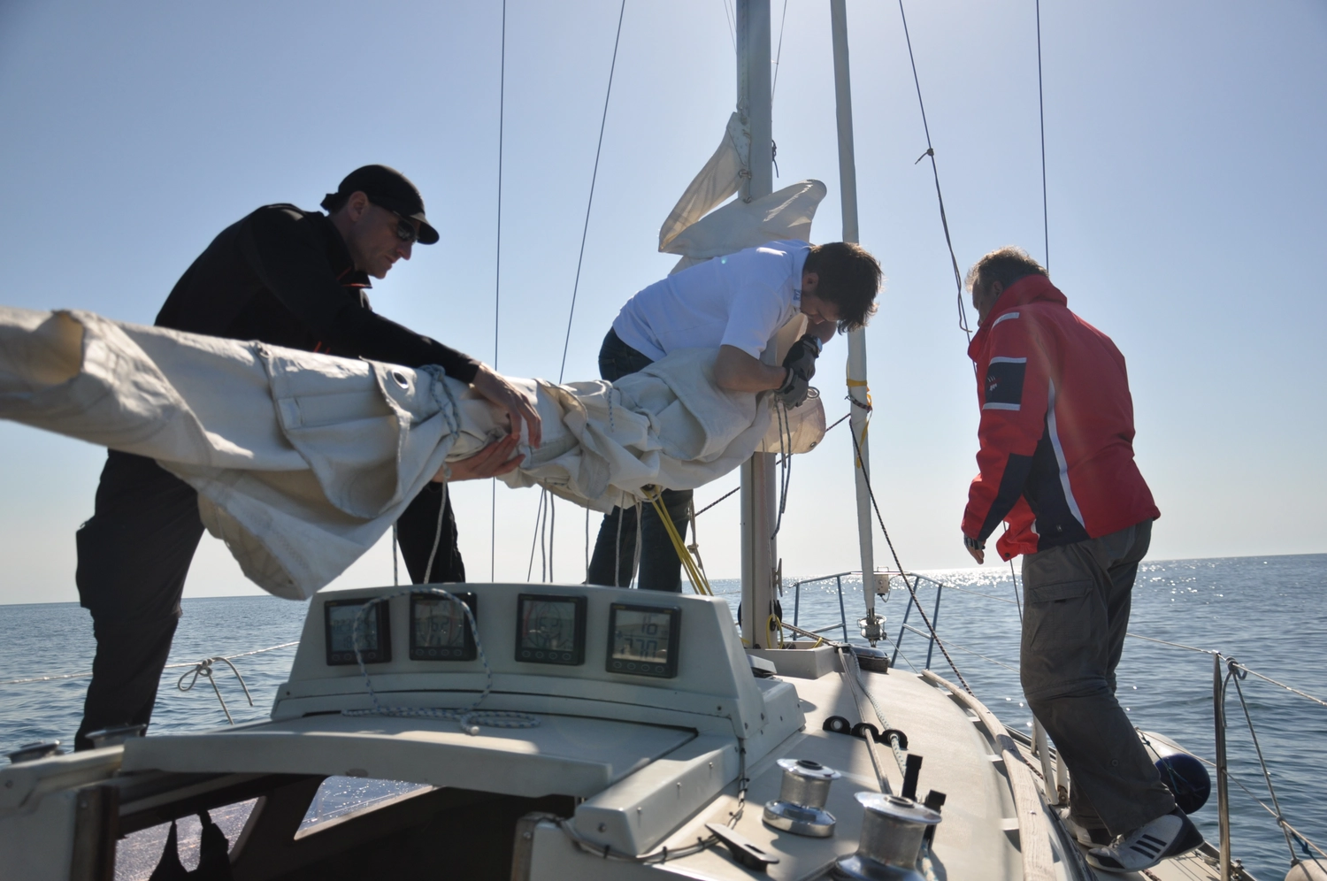 Motorboot-u. Segelschule Ravensburg - Wassersportausbildung und lizenzierung - Drei Personen arbeiten auf einem Segelboot zusammen, um ein großes weißes Segel auf dem Wasser zu bedienen.