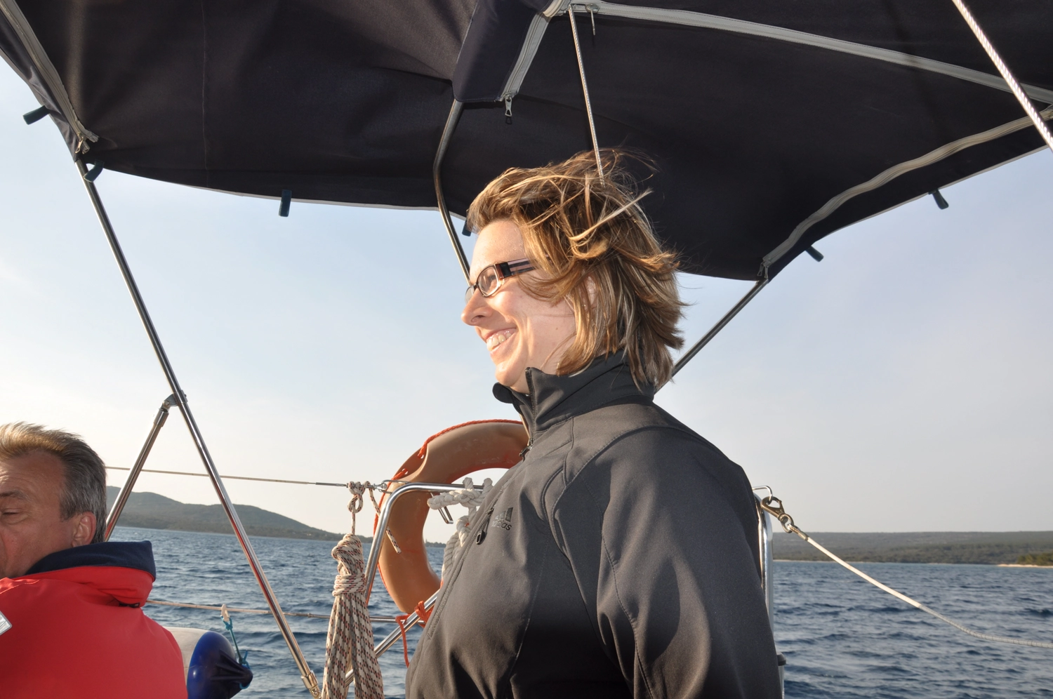 Motorboot-u. Segelschule Ravensburg - Wassersportausbildung und lizenzierung - Eine Frau lächelt, während sie am Steuer eines Segelboots unter einem schwarzen Sonnenschirm steht.