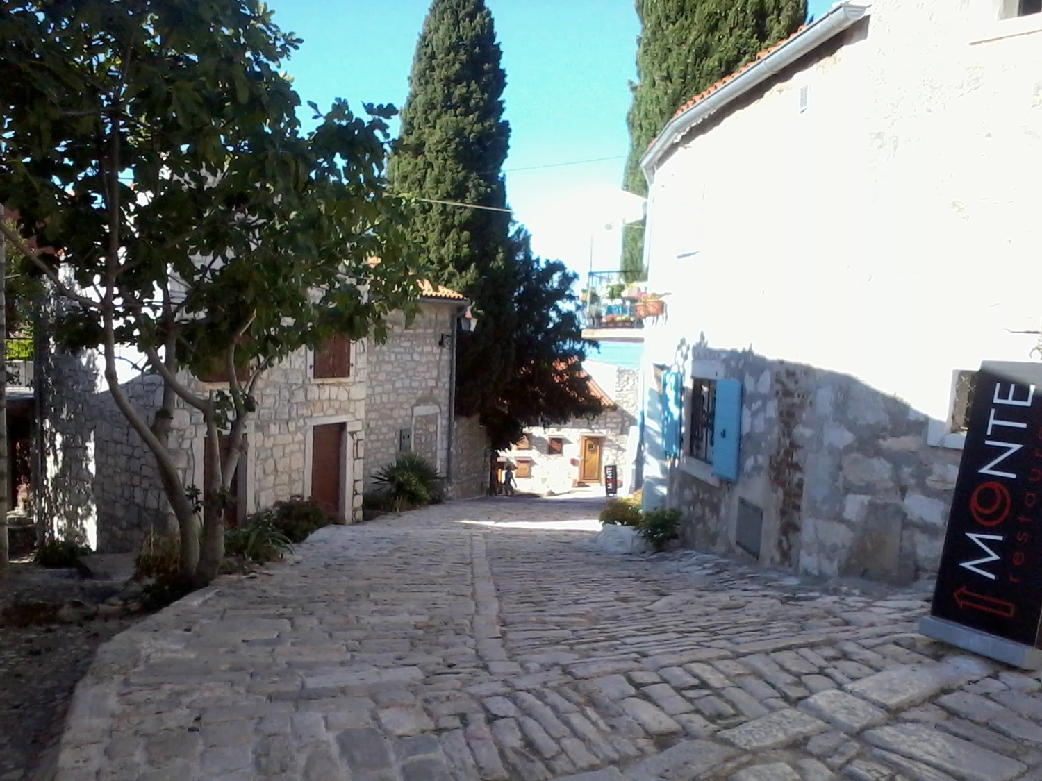 Motorboot-u. Segelschule Ravensburg - Wassersportausbildung und lizenzierung - Ein schmaler, sonnenbeschienener Steinweg führt zwischen charmanten alten Gebäuden mit weißen Mauern und grünen Bäumen hindurch.