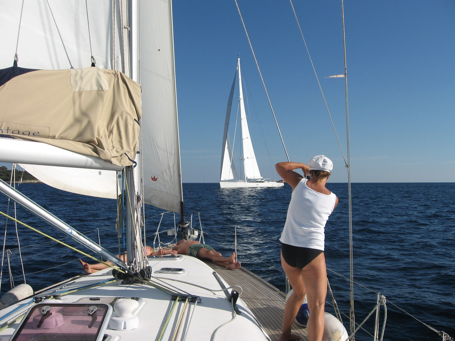 Motorboot-u. Segelschule Ravensburg - Wassersportausbildung und lizenzierung - Eine Person in Weiß steht auf dem Deck eines Segelboots und blickt auf ein anderes weißes Segelboot in der Ferne auf einem klaren blauen Meer.