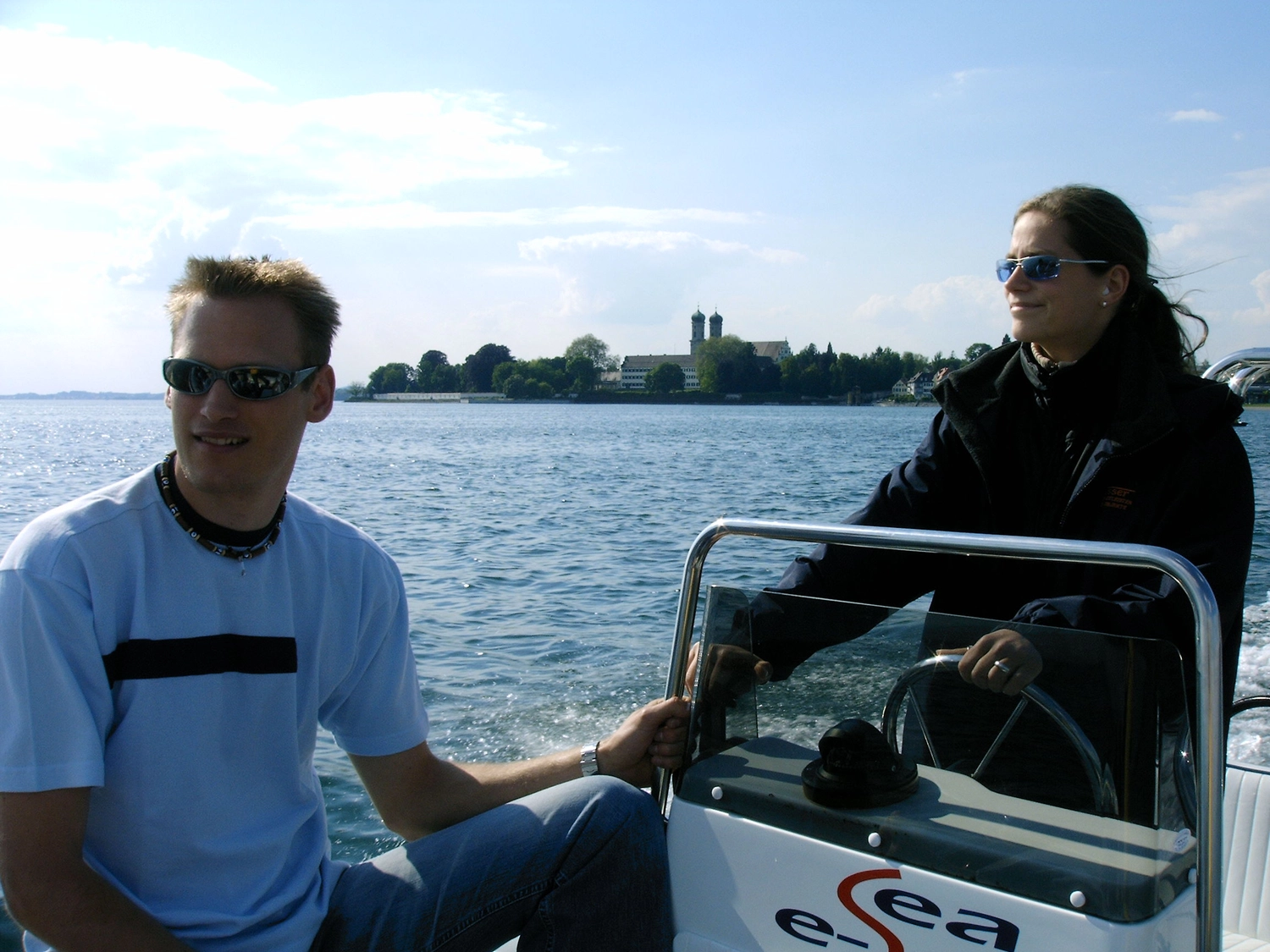 Motorboot-u. Segelschule Ravensburg - Wassersportausbildung und lizenzierung - Ein Mann in einem blauen Hemd und eine Frau in einer schwarzen Jacke sitzen an einem sonnigen Tag am Steuer eines kleinen Motorboots.