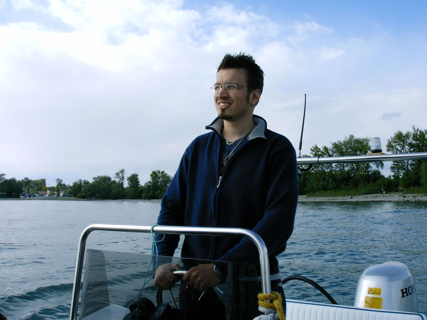 Motorboot-u. Segelschule Ravensburg - Wassersportausbildung und lizenzierung - Ein Mann in einer dunklen Jacke steht auf einem Boot und blickt auf die Küste in der Ferne.