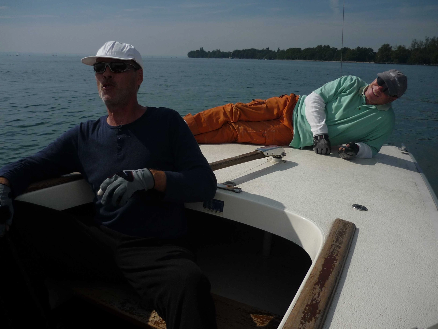 Motorboot-u. Segelschule Ravensburg - Wassersportausbildung und lizenzierung - Ein Mann in einem dunklen, langärmeligen Hemd steuert ein Segelboot, während eine andere Person in einem grünen Hemd und einer orangefarbenen Hose auf dem Deck entspannt.