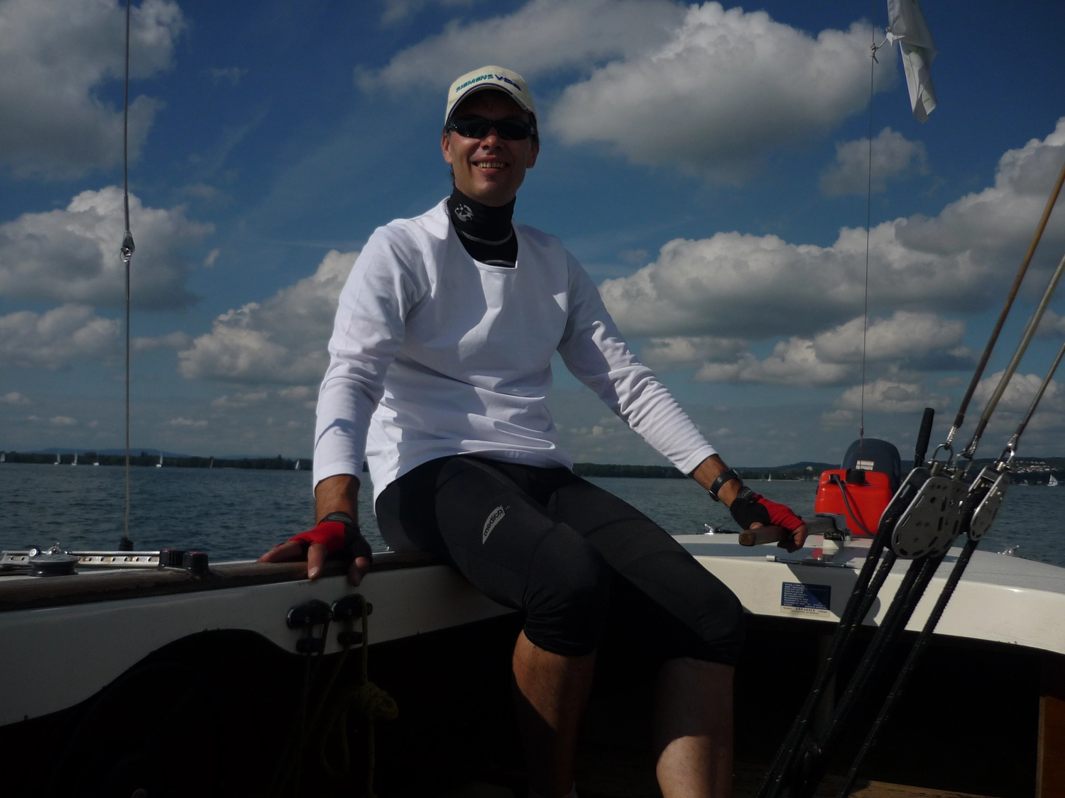 Motorboot-u. Segelschule Ravensburg - Wassersportausbildung und lizenzierung - Ein Mann in einem weißen Langarmhemd und einem weißen Hut sitzt unter einem hellen, bewölkten Himmel am Rand eines Segelboots.