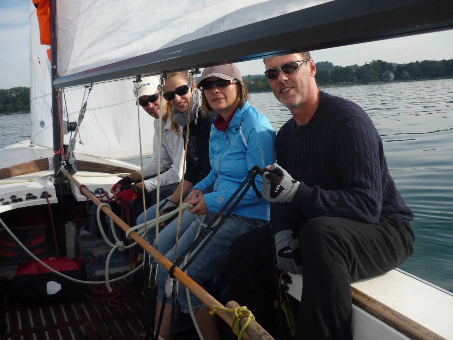 Motorboot-u. Segelschule Ravensburg - Wassersportausbildung und lizenzierung - Vier Personen sitzen lächelnd an der Reling eines Segelboots, während sie über das Wasser gleiten.