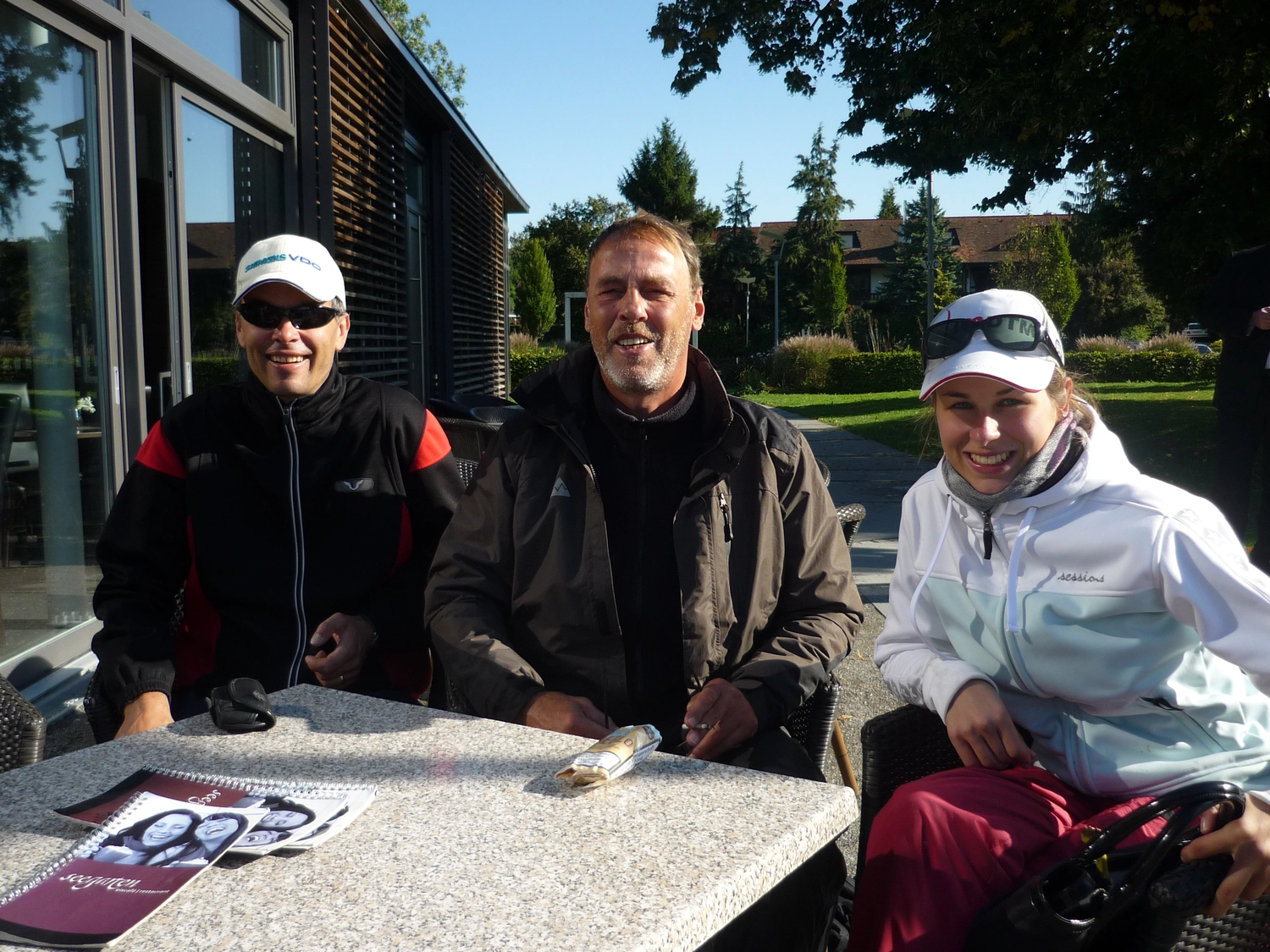 Motorboot-u. Segelschule Ravensburg - Wassersportausbildung und lizenzierung - Drei Männer in lässigen Jacken sitzen an einem sonnigen Tag um einen Tisch im Freien.
