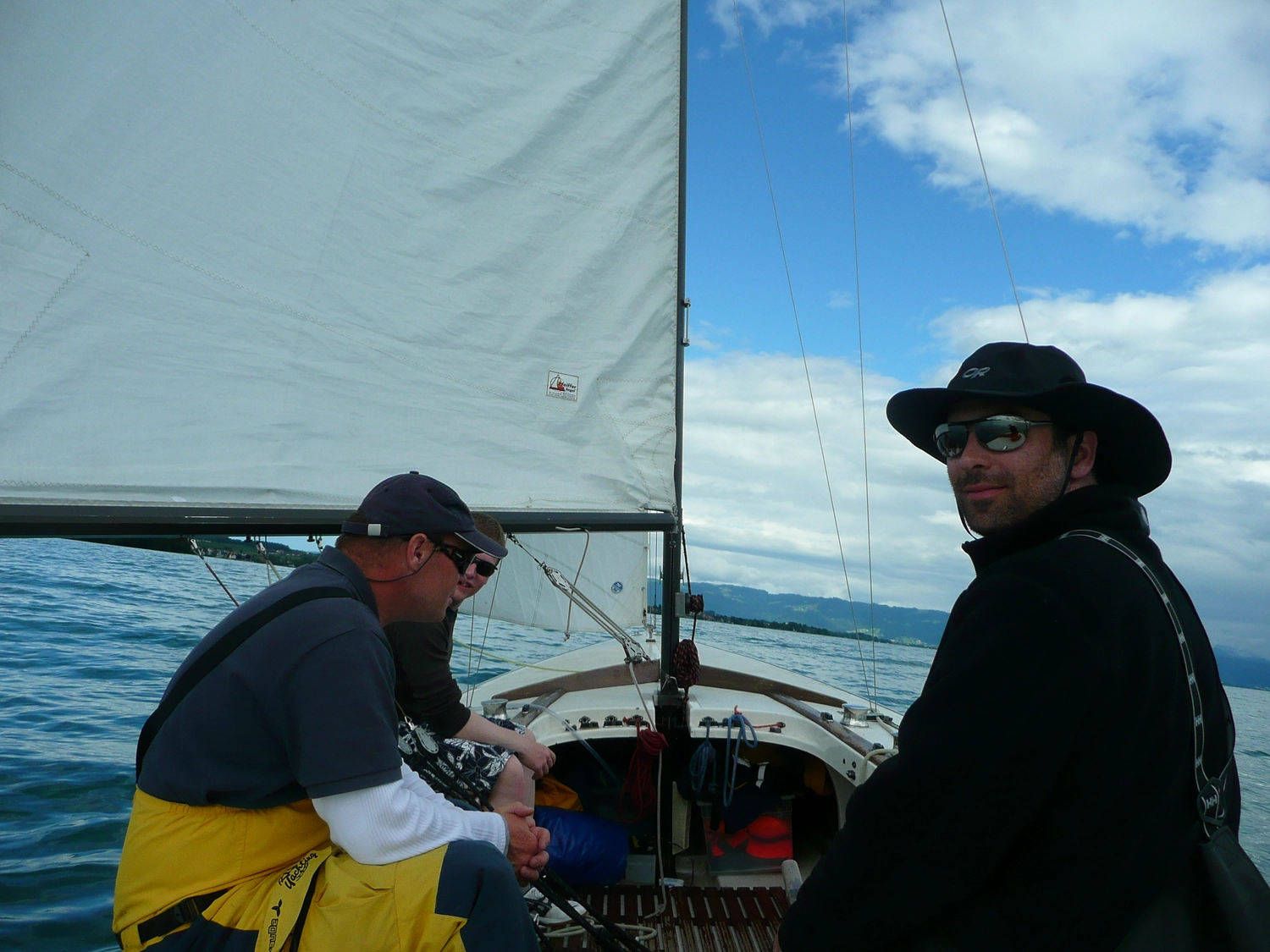 Motorboot-u. Segelschule Ravensburg - Wassersportausbildung und lizenzierung - Ein Mann in einem dunklen Polohemd und einer weißen Kappe sitzt auf einem Segelboot und blickt zurück zum Steuerstand.