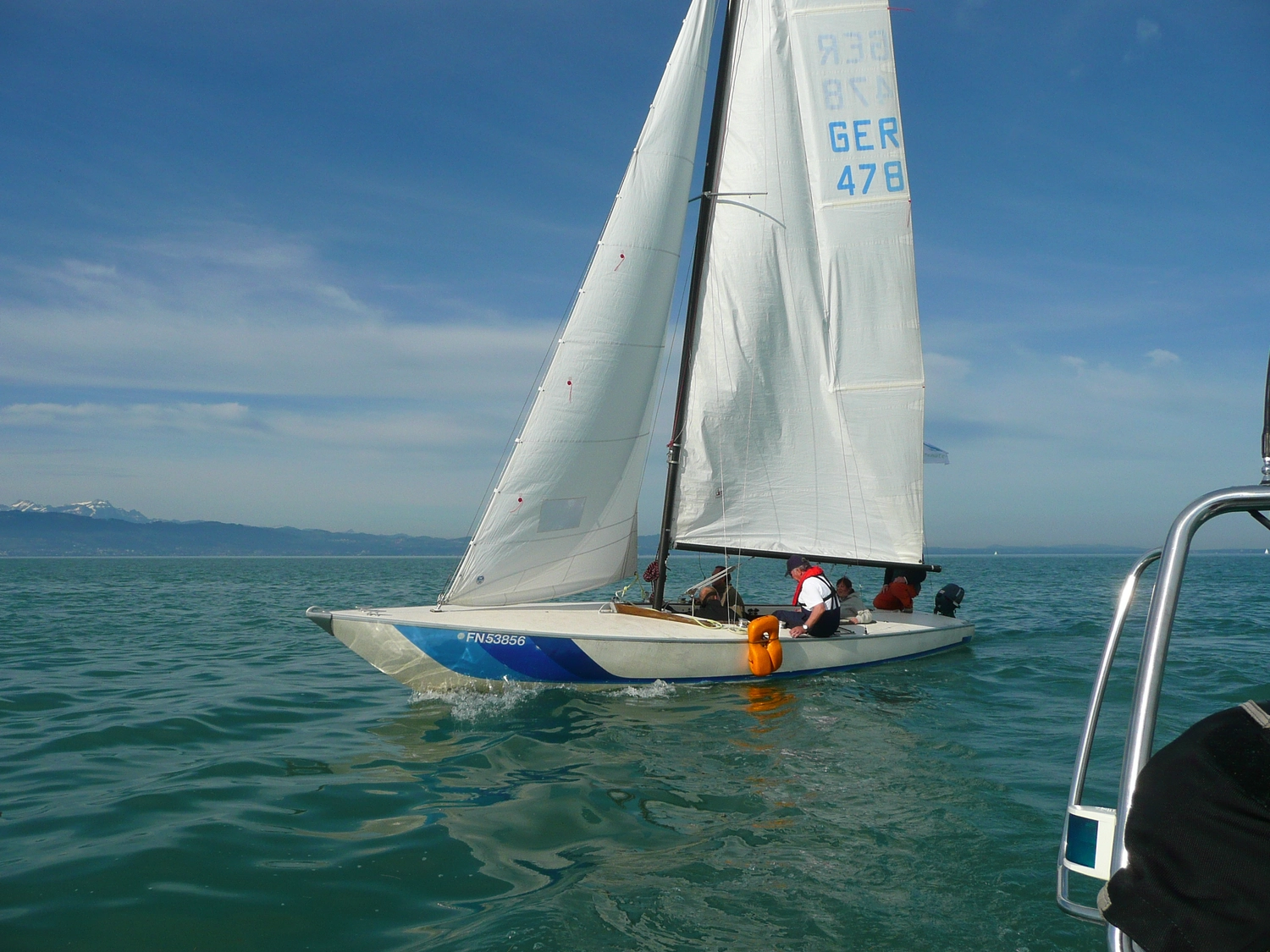 Motorboot-u. Segelschule Ravensburg - Wassersportausbildung und lizenzierung - Ein weißes Segelboot mit blauen Akzenten segelt auf ruhigem, blauem Wasser unter strahlend blauem Himmel.