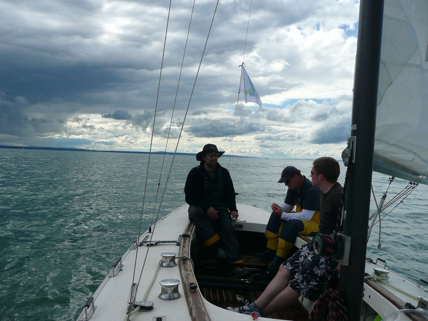 Motorboot-u. Segelschule Ravensburg - Wassersportausbildung und lizenzierung - Eine Gruppe von Menschen sitzt unter bewölktem Himmel auf einem Segelboot und blickt aufs Wasser hinaus.