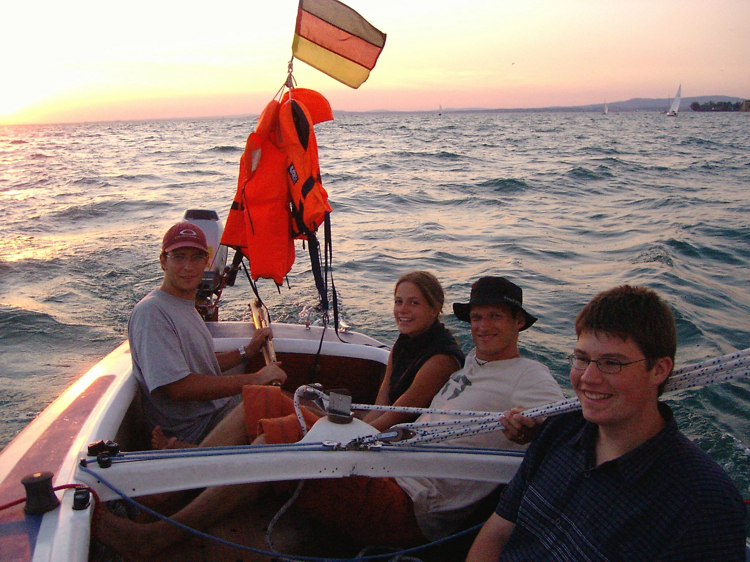 Motorboot-u. Segelschule Ravensburg - Wassersportausbildung und lizenzierung - Eine Gruppe von Menschen entspannt sich bei Sonnenuntergang auf einem Segelboot, unter dem eine kleine Flagge weht.