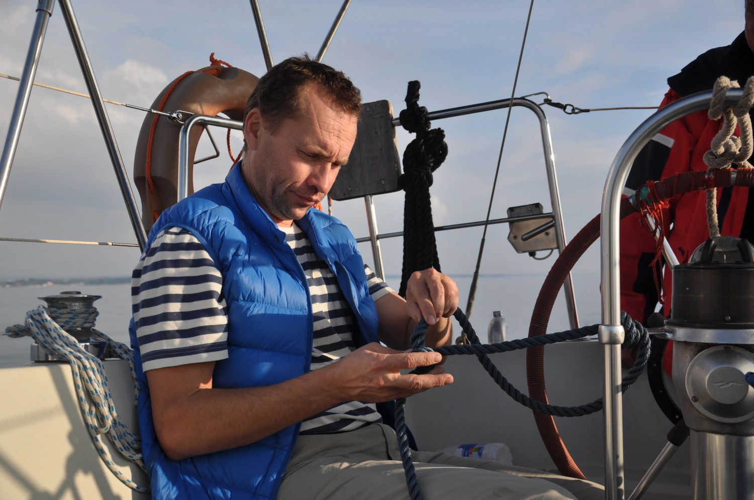Motorboot-u. Segelschule Ravensburg - Wassersportausbildung und lizenzierung - Ein Mann in blauer Weste und gestreiftem Hemd hantiert vorsichtig mit den Takelageleinen an Deck eines Segelboots.