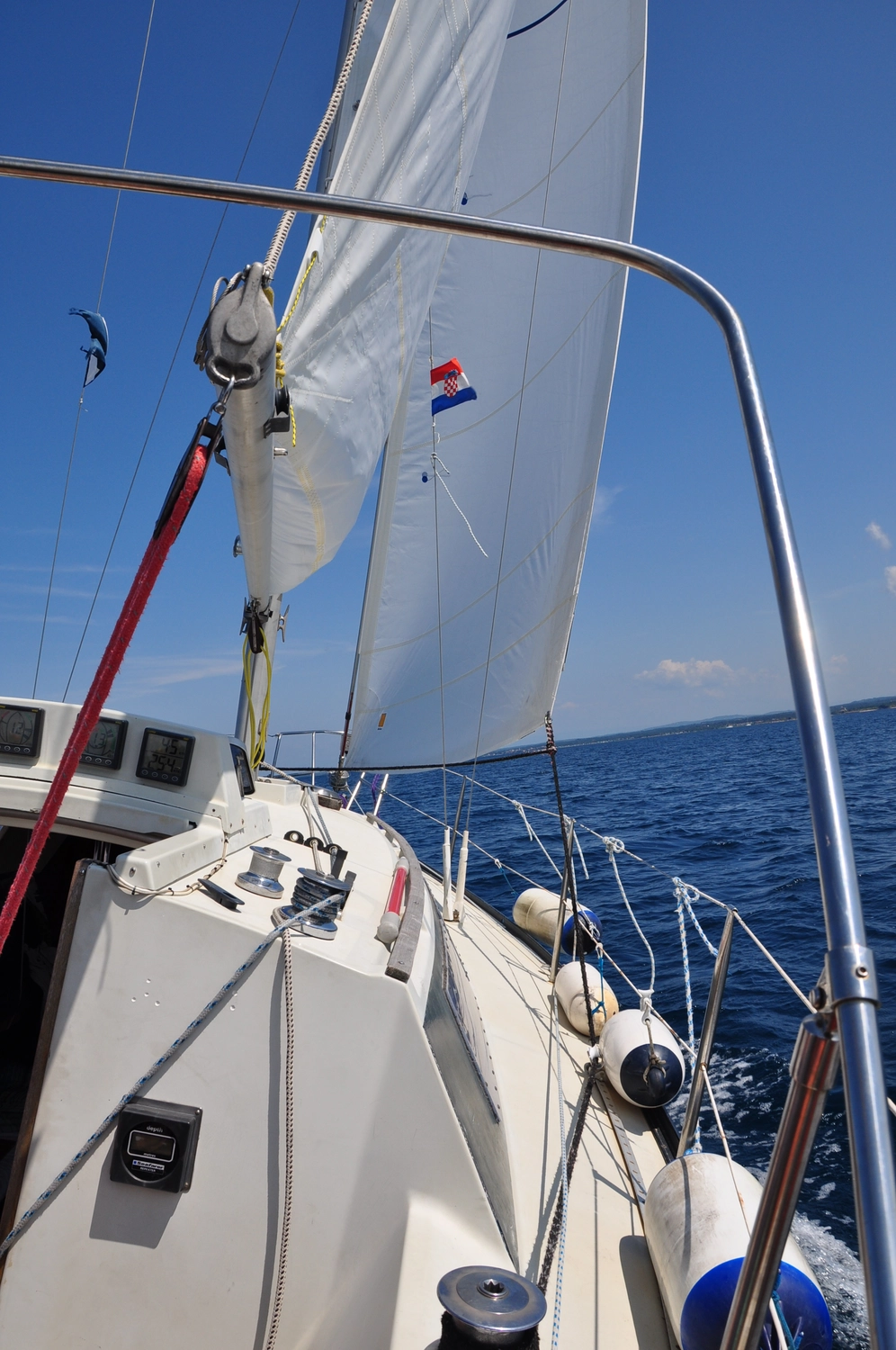 Motorboot-u. Segelschule Ravensburg - Wassersportausbildung und lizenzierung - Blick vom Deck auf ein weißes Segelboot, das unter klarem Himmel durch tiefblaues offenes Wasser gleitet; es sind getakelte Segel gesetzt und mehrere weiße Fender hängen an der Seite.