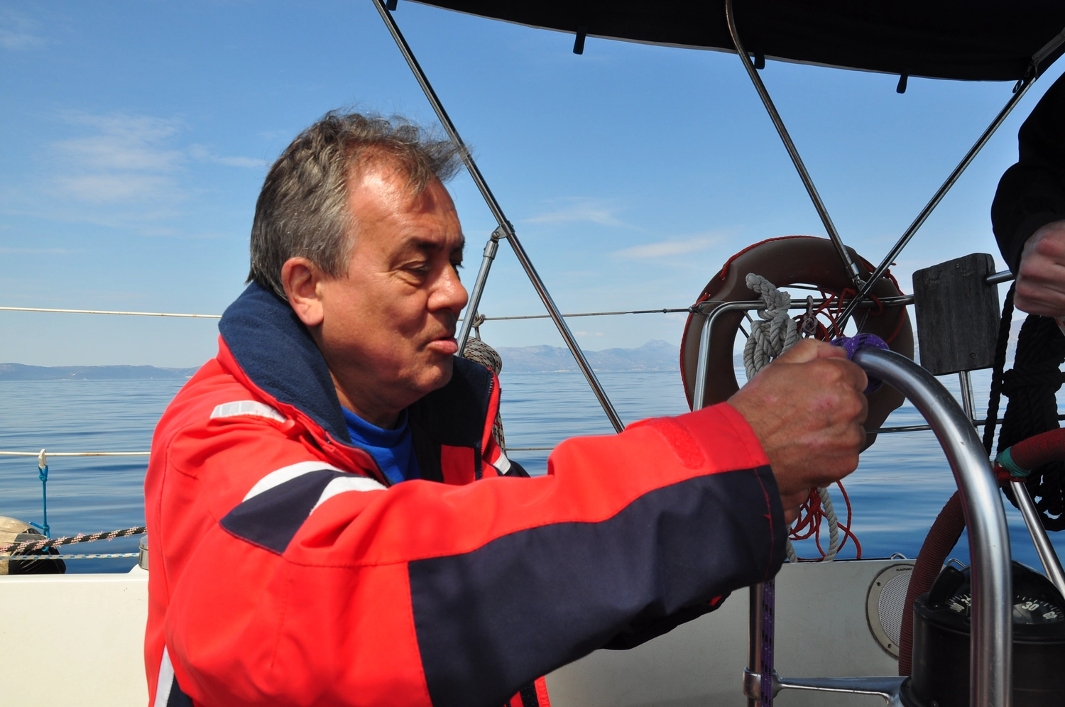 Motorboot-u. Segelschule Ravensburg - Wassersportausbildung und lizenzierung - Ein Mann in einer rot-blauen Segeljacke steuert ein Boot mit einem Metallsteuerrad.