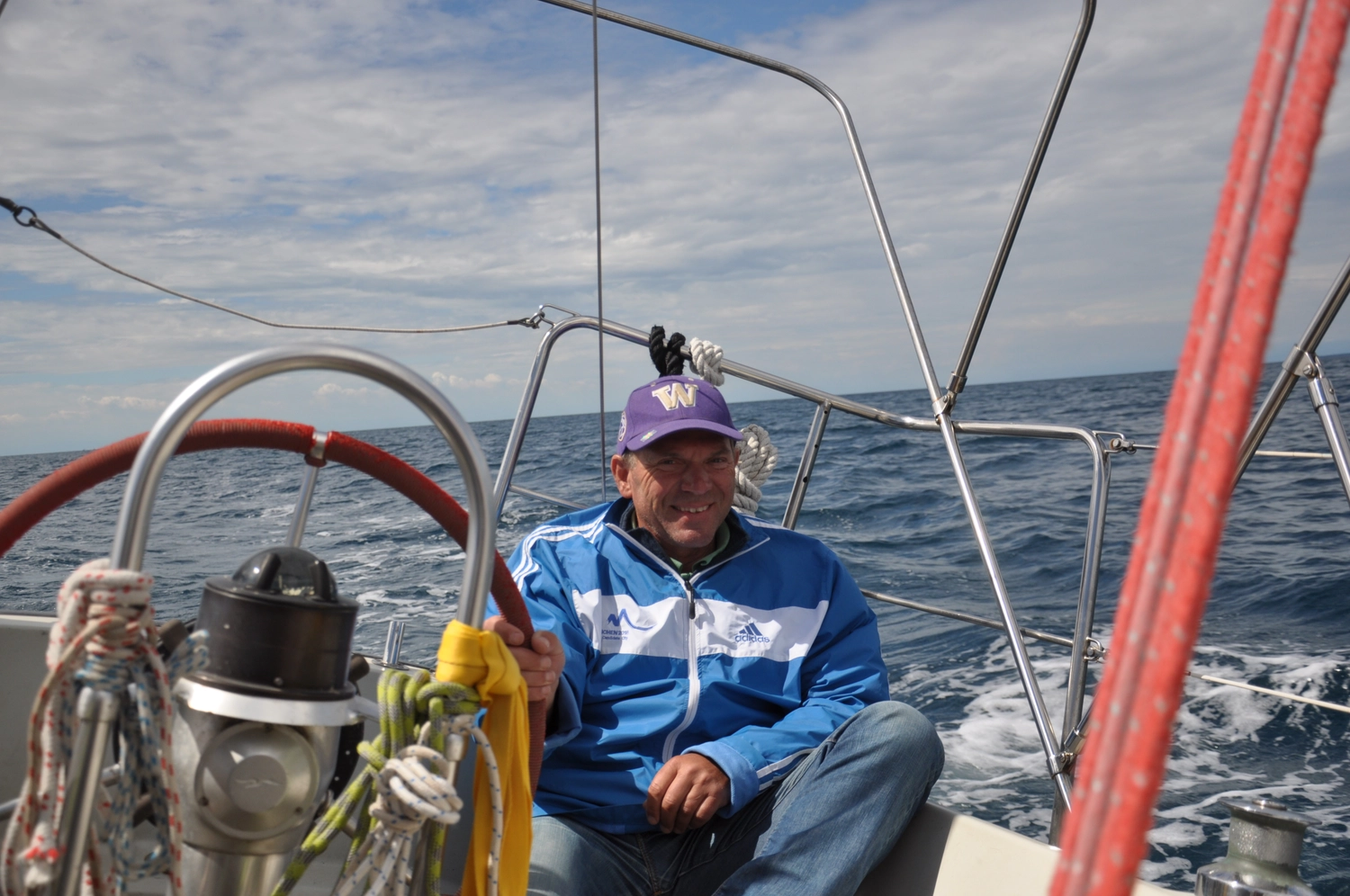 Motorboot-u. Segelschule Ravensburg - Wassersportausbildung und lizenzierung - Ein Mann mit lila Baseballkappe und blauer Jacke lächelt aus dem Cockpit eines Segelboots.