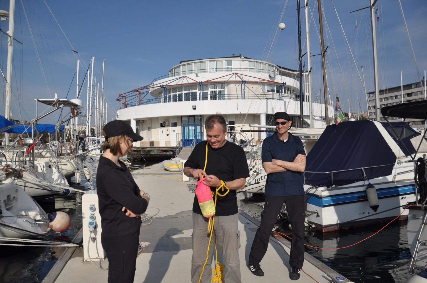 Motorboot-u. Segelschule Ravensburg - Wassersportausbildung und lizenzierung - Drei Personen stehen auf einem Steg neben einem Boot, im Hintergrund ist ein großes Gebäude zu sehen. Sie werben für einen kombinierten Segel- und Motorbootkurs.
